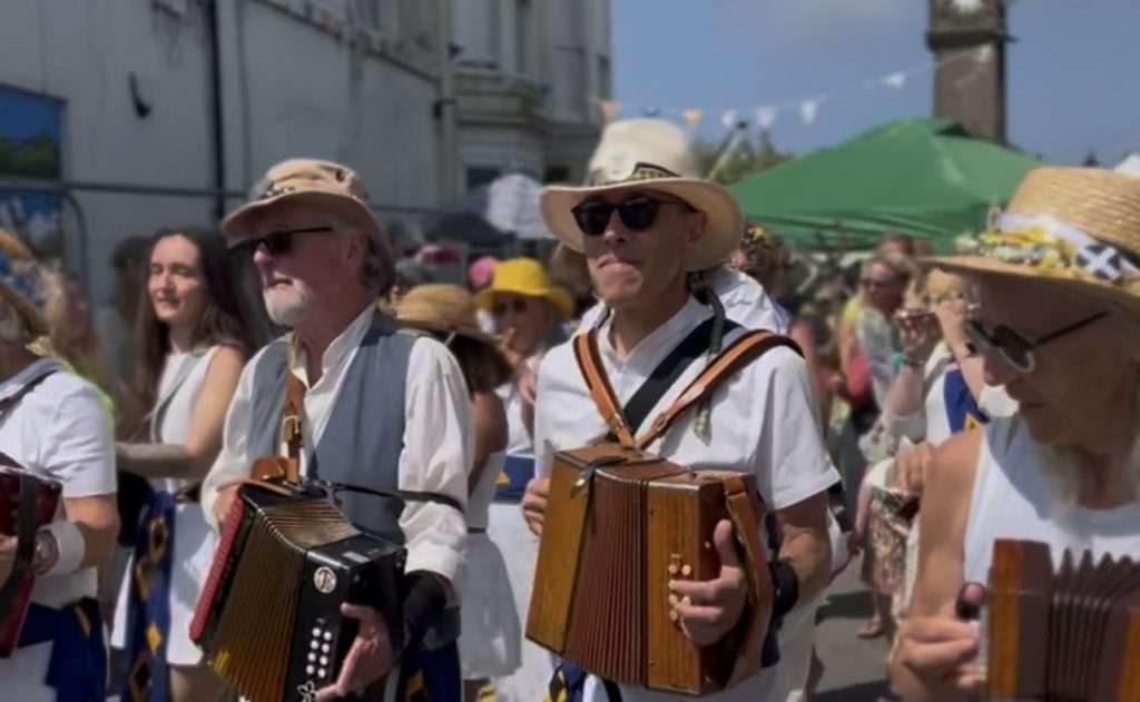 Musicians playing accordions in a daytime Lafrowda Festival street procession. The performers wear hats, sunglasses and traditional-style outfits as they move through a busy street lined with festivalgoers and bunting.
